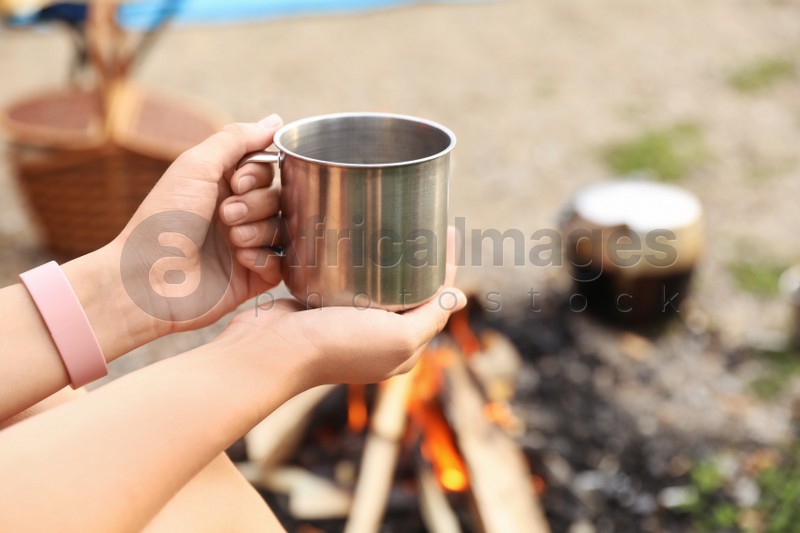 Young woman with mug near bonfire outdoors, focus on hands. Camping season