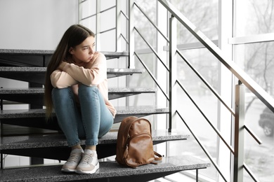 Upset teenage girl with backpack sitting on stairs indoors. Space for text