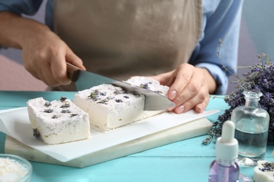 Woman cutting hand made soap bar with lavender flowers at light blue wooden table, closeup