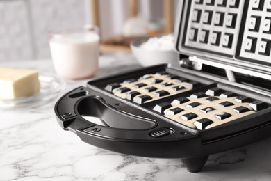 Photo of Belgian waffle maker with dough on white marble table, closeup