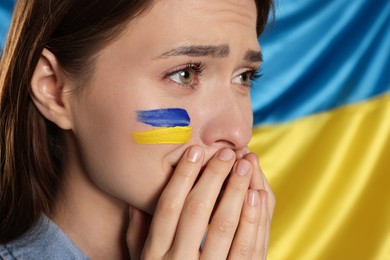 Photo of Sad young woman with clasped hands near Ukrainian flag, closeup