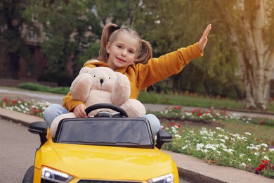 Photo of Cute little girl with toy bear driving children's car in park