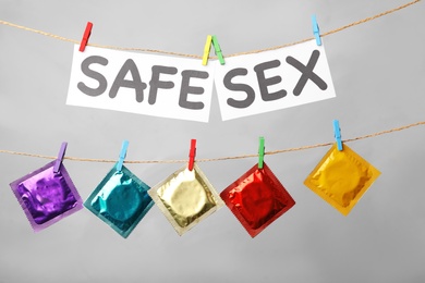 Paper sheets with words SAFE SEX and colorful condoms hanging on clothesline against light grey background