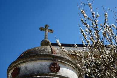 Roof of old chapel with stone cross near blossoming tree against blue sky, closeup