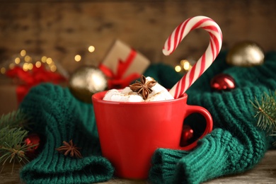 Cup of tasty cocoa with marshmallows and Christmas candy cane on wooden table against blurred festive lights