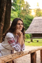 Photo of Beautiful woman wearing embroidered shirt near wooden railing in countryside. Ukrainian national clothes