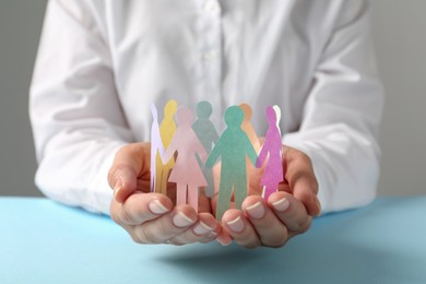 Photo of Woman holding paper human figures at light blue table against grey background, closeup. Diversity and inclusion concept
