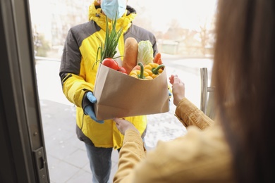 Courier in medical mask giving paper bag with groceries to woman at doorway, closeup. Delivery service during Covid-19 quarantine