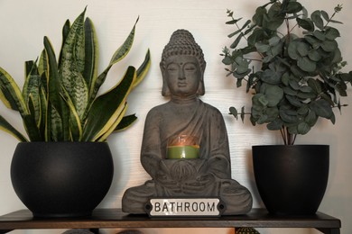 Buddha sculpture with candle and green houseplants on shelf indoors. Interior design