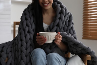 Woman with chunky knit blanket and cup in armchair at home, closeup