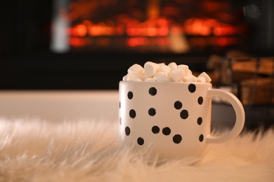 Cup of hot drink with marshmallows on fluffy rug near fireplace at home. Space for text