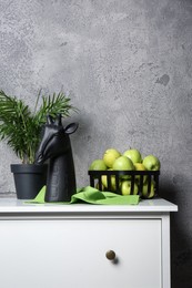 Photo of Stylish decor, basket with apples and houseplant on chest of drawers near grey wall indoors, space for text. Interior design