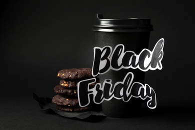 Takeaway paper coffee cup with phrase Black Friday and cookies against dark background