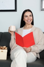 Photo of Happy woman reading greeting card while drinking coffee on sofa in living room