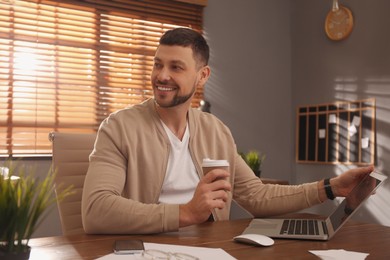 Freelancer with cup of coffee working on laptop at table indoors