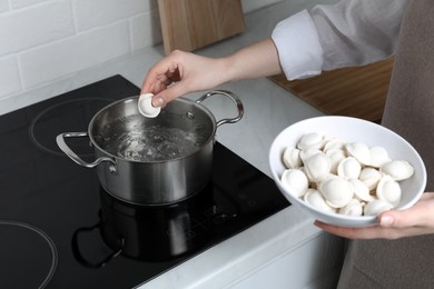 Woman putting frozen dumplings into saucepan with boiling water on cooktop in kitchen, closeup