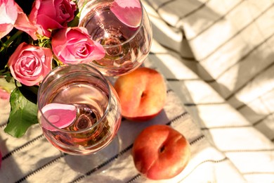 Photo of Glasses of delicious rose wine with petals, flowers and peaches on white picnic blanket outside, above view. Space for text