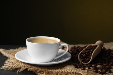 Cup of hot aromatic coffee and scoop with roasted beans on black table against dark background
