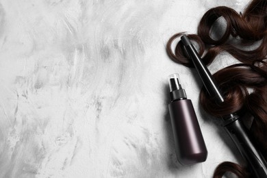 Photo of Spray bottle with thermal protection, lock of brown hair and modern curling iron on grey table, flat lay. Space for text