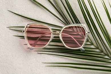 Stylish sunglasses and palm leaf on sand, top view