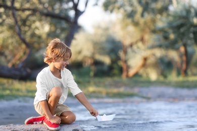 Photo of Cute little boy with paper ship at river, space for text. Child spending time in nature