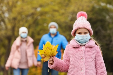 Cute girl in medical mask walking with her parents outdoors on autumn day. Protective measures during coronavirus quarantine