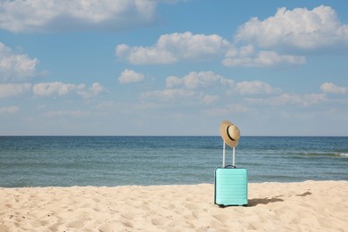 Turquoise suitcase with straw hat on sandy beach near sea, space for text