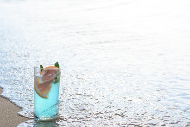Glass of refreshing drink with grapefruit and mint getting hit by wave on sea beach, space for text
