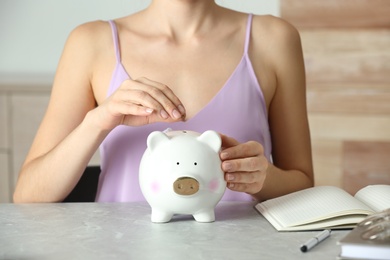 Woman putting coin into piggy bank at grey marble table, closeup. Money savings