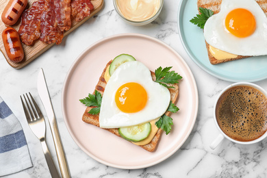 Tasty sandwiches with heart shaped fried eggs for romantic breakfast on white marble table, flat lay