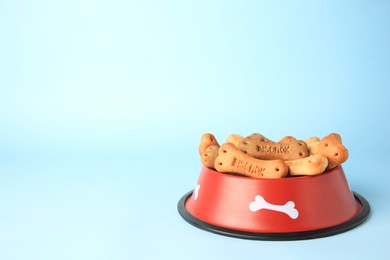 Photo of Bone shaped dog cookies in feeding bowl on light blue background, space for text