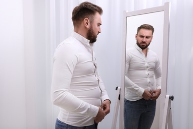 Upset man wearing tight shirt in front of mirror at home. Overweight problem