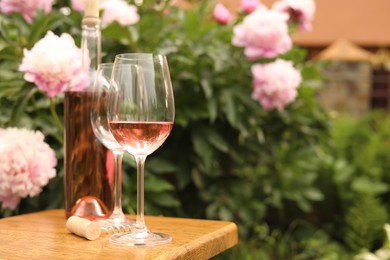 Glasses and bottle with rose wine on wooden table near beautiful peonies, space for text