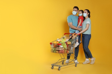 Family with protective masks and shopping cart full of groceries on yellow background. Space for text