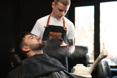 Professional hairdresser with shaving foam near bearded client in barbershop