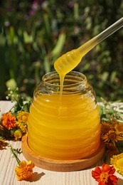 Taking delicious fresh honey with dipper from glass jar surrounded by beautiful flowers on table in garden