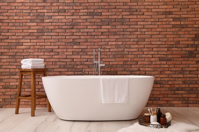 Modern ceramic bathtub and tray with toiletries near brick wall indoors