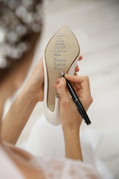 Young bride writing her single friends names on shoe indoors, closeup. Wedding superstition