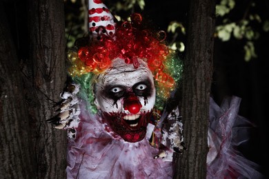Terrifying clown hiding behind trees outdoors at night. Halloween party costume