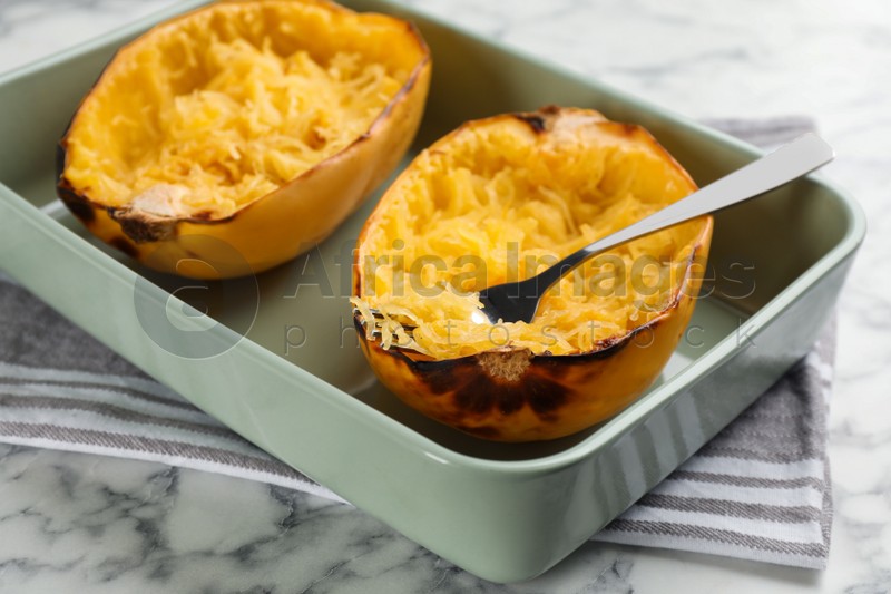 Halves of cooked spaghetti squash and fork in baking dish on white marble table, closeup