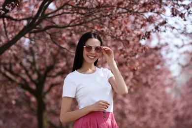 Pretty young woman with sunglasses near beautiful blossoming trees outdoors. Stylish spring look