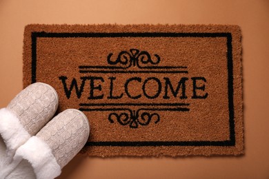 Photo of Doormat with word Welcome and slippers on brown background, flat lay