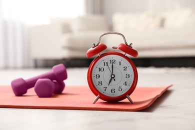 Alarm clock, yoga mat and dumbbells on wooden floor indoors. Morning exercise