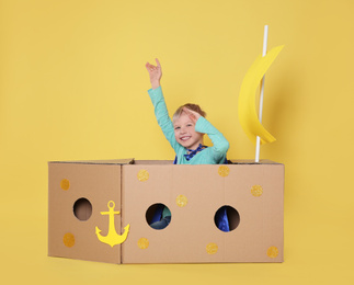 Little child playing with ship made of cardboard box on yellow background
