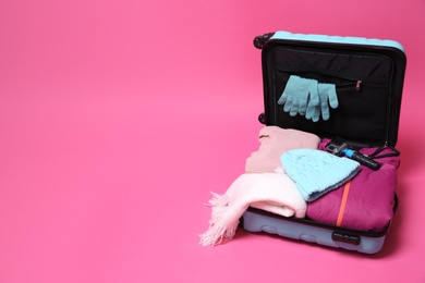 Open suitcase with warm clothes and action camera on color background, space for text. Winter vacation