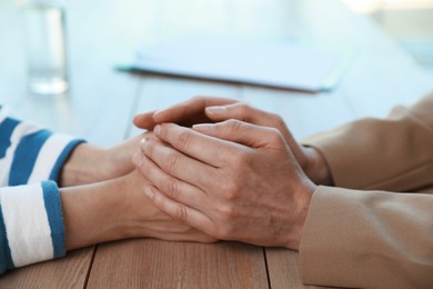 Psychotherapist holding patient's hands at table indoors, closeup