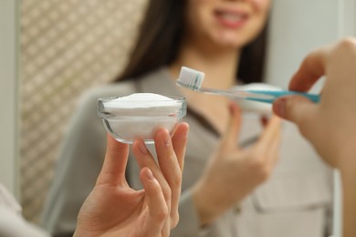Photo of Young woman with toothbrush and bowl of baking soda near mirror indoors, closeup