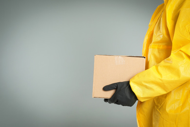 Man in chemical protective suit holding cardboard box on light grey background, closeup view with space for text. Prevention of virus spread