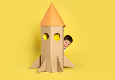 Photo of Cute little child playing with cardboard rocket on yellow background
