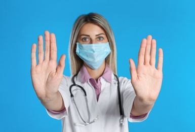 Doctor in protective mask showing stop gesture on light blue background. Prevent spreading of coronavirus